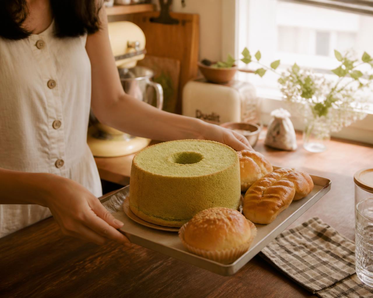 Home baker carrying a tray of pandan chiffon cake and buns in her kitchen